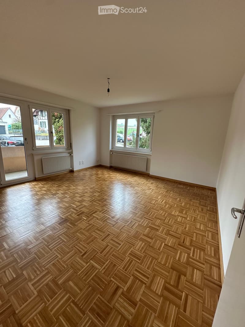 empty room, white walls, wooden parquet floor, 2 sliding doors with windows, wall radiators, light bulb hanging from ceiling