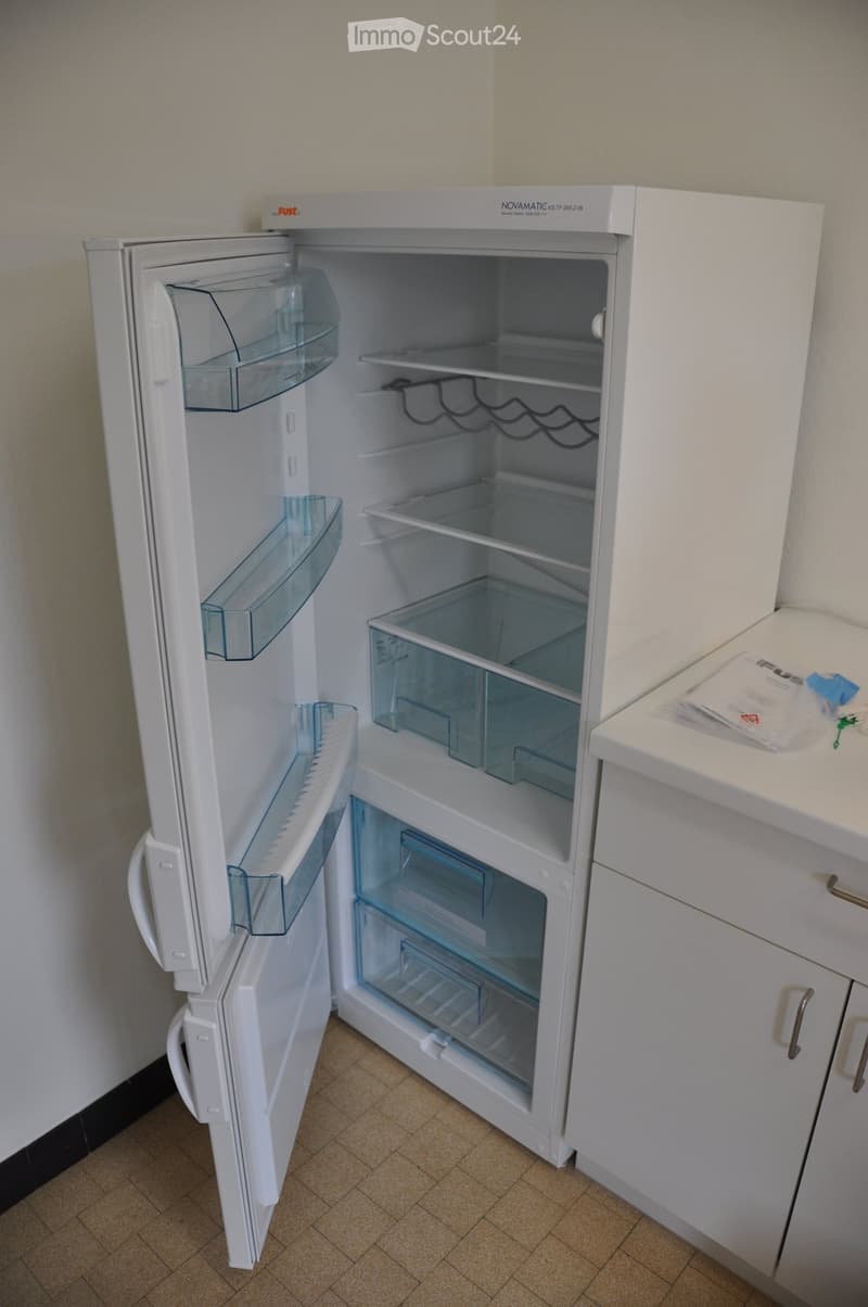 Kitchen. A white refrigerator with multiple compartments is open, showing empty shelves and drawers. The kitchen features light-colored cabinets and a tiled floor.