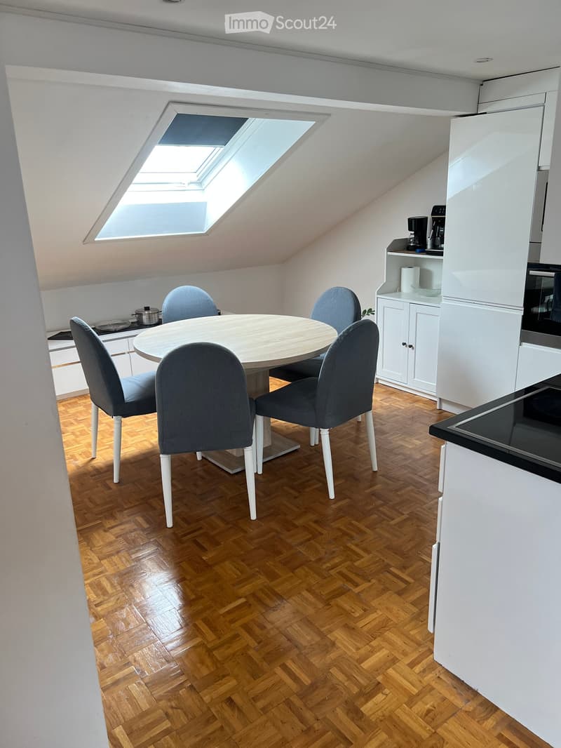 Furnished Room. A modern, compact kitchen and dining area with white cabinets, a round wooden dining table, four grey upholstered chairs, and a small kitchen counter with a coffee maker and other appliances.