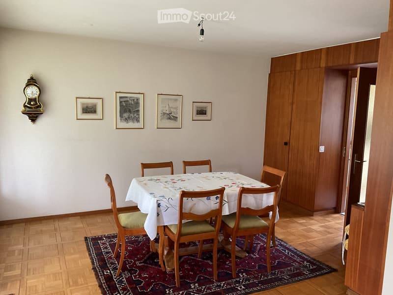 Dining room with wooden floor, dining table with floral tablecloth, six wooden chairs, wall-mounted clock, and several framed pictures on the wall.
