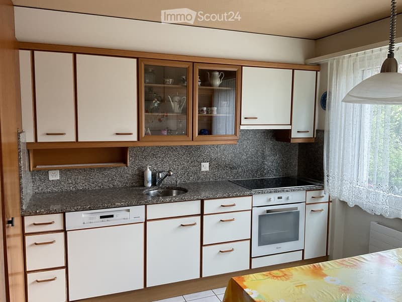 A modern kitchen with white cabinets, granite countertops, a sink, an oven, and a dishwasher. A table with a floral tablecloth is partially visible.