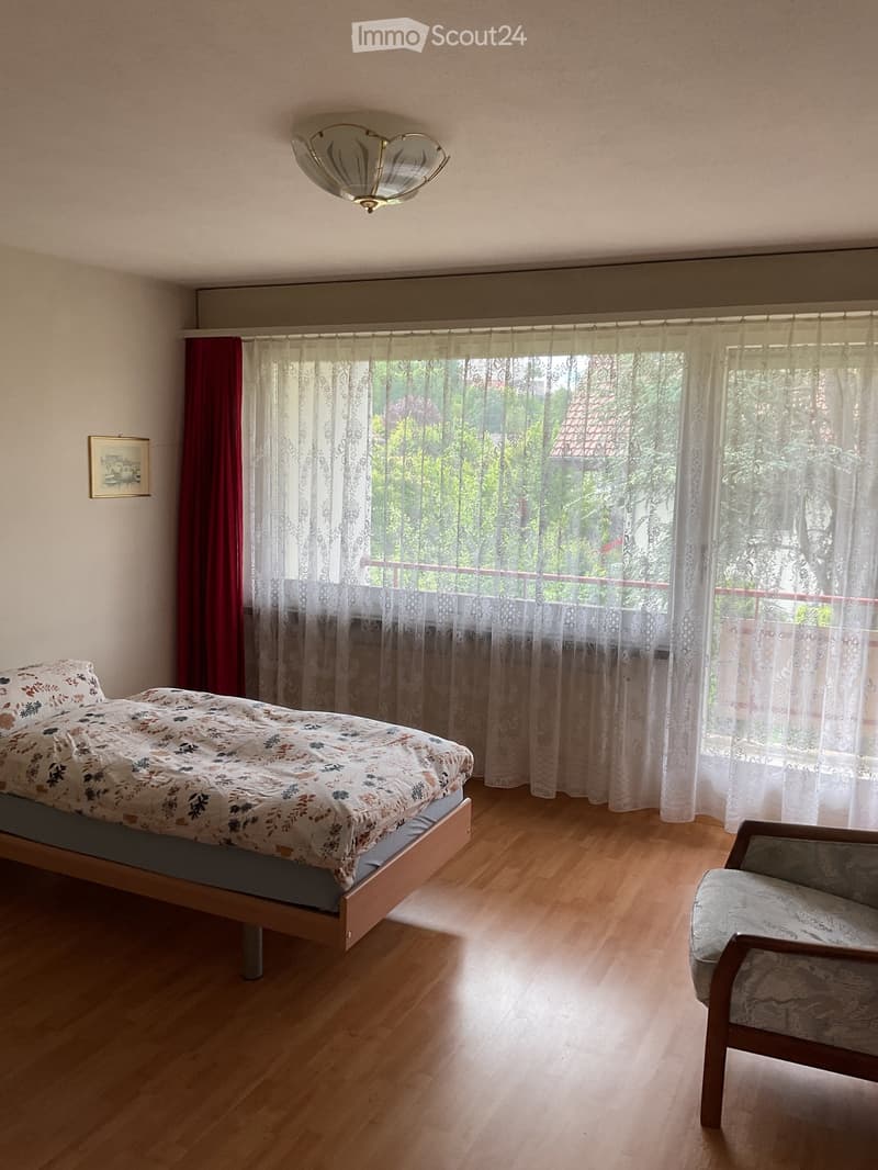 Bedroom with wooden floor, large window with white and red curtains, bed with floral duvet, armchair, and picture frame on wall.