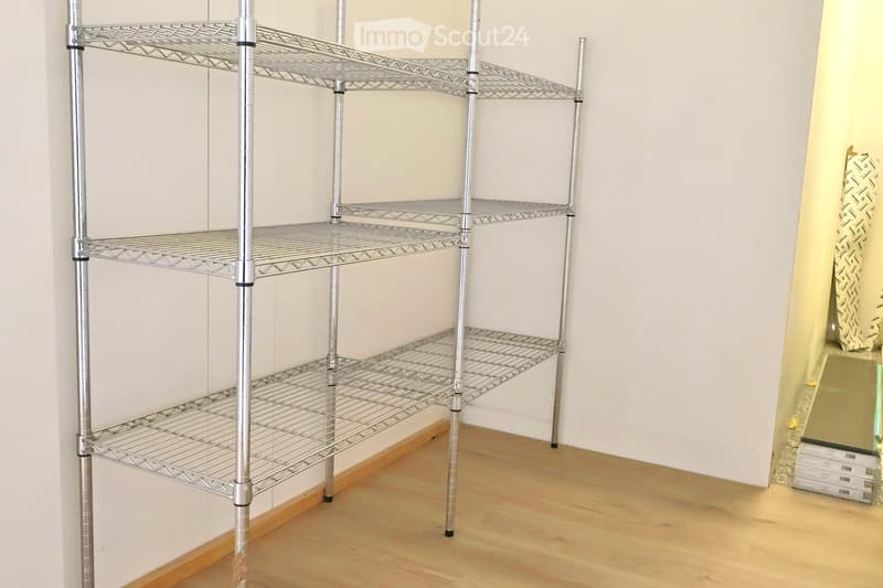 An empty room with a white wall and a metal shelving unit against it. The floor is wooden.