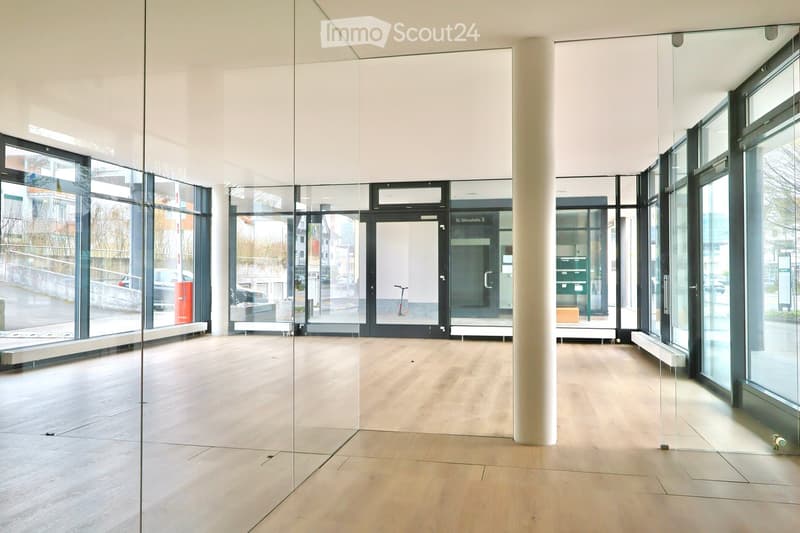 An empty room with glass walls, a white ceiling, and a wooden floor. There are glass doors and windows, and a white pillar in the center. Outside, there are cars parked and a building with glass windows.