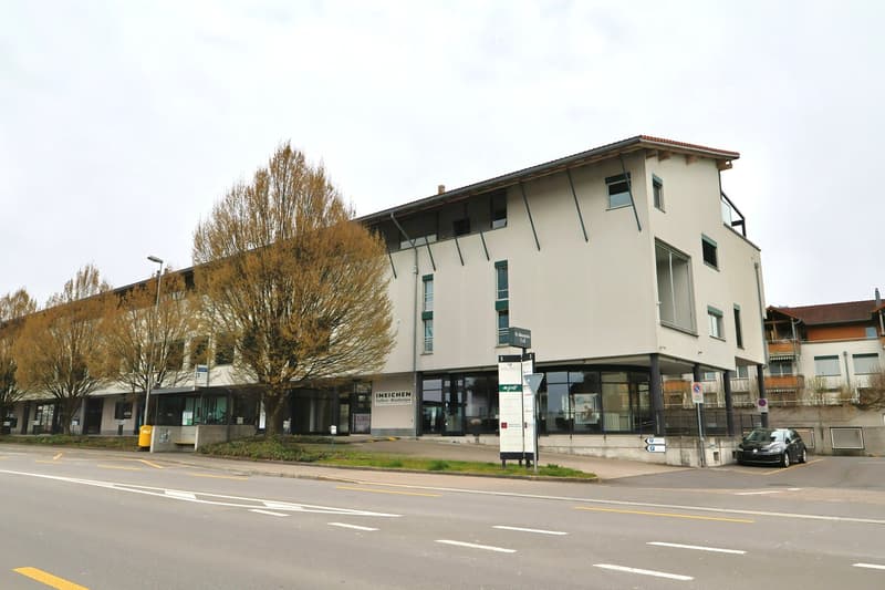 A modern building with glass windows and a red roof, situated on a street with trees and a parked car.