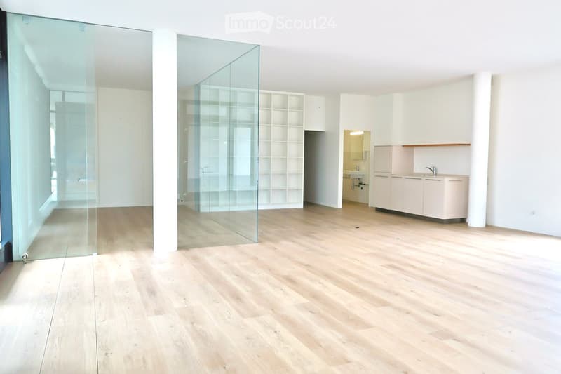 An empty room with a glass wall, wooden floor, and white walls. There is a white cabinet with a sink and faucet in the corner.