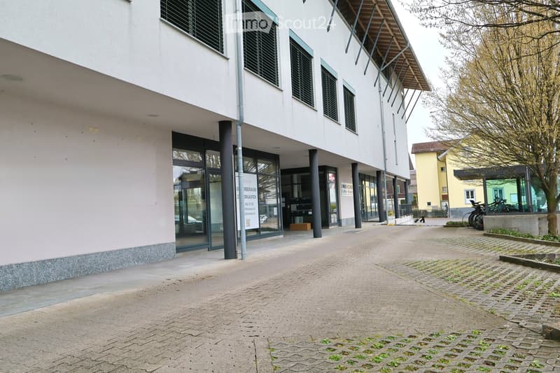 Modern building with glass windows and a signboard. The entrance is flanked by two pillars. A person is walking in the distance. A tree and bicycles are parked nearby.