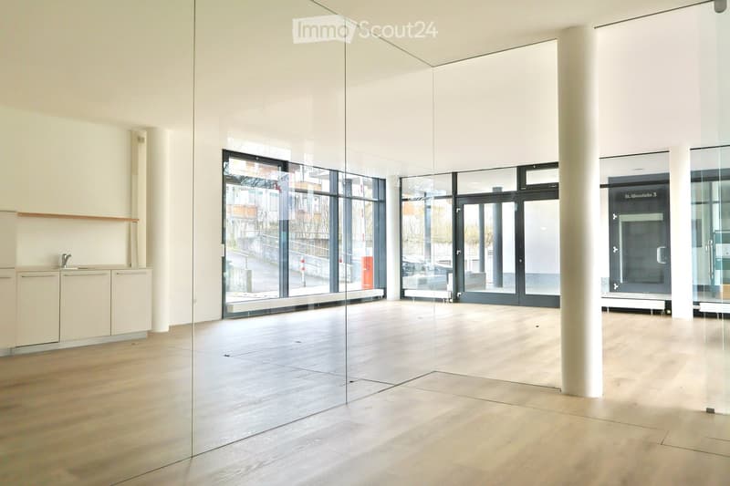 An empty room with glass walls, a white ceiling, and a wooden floor. There are white cabinets and a sink in the left corner, and a pillar in the center.