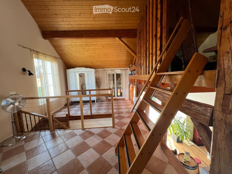 Spacious hallway with wooden ceiling, tiled floor, and large windows. Features a staircase leading to an upper level, a fan on the left, and a view into an adjacent room with a white wardrobe and a bed.