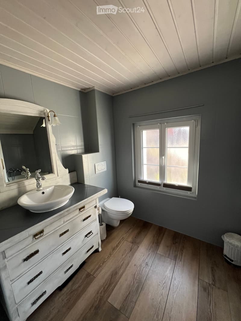 Modern bathroom with a white sink, mirror, and toilet, featuring wooden flooring and grey walls.