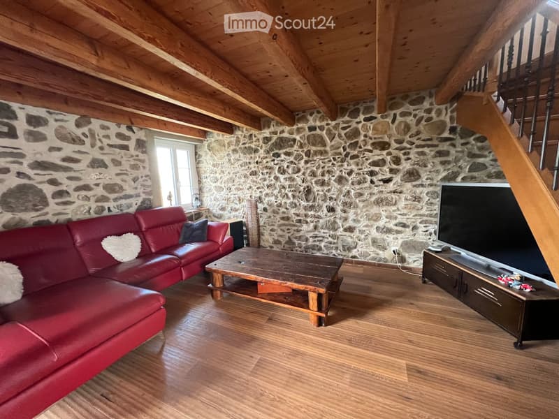 Living room with exposed wooden beams, stone walls, red leather sofa, wooden coffee table, and TV on a stand.