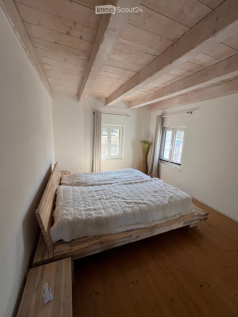 Cozy bedroom with wooden bed frame, fluffy white bedding, wooden nightstand, and large windows with curtains.