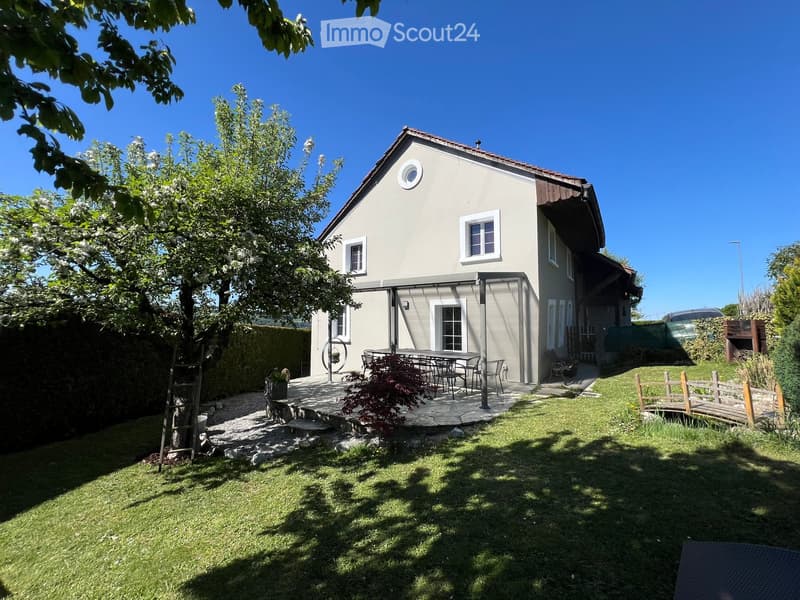 A well-maintained garden with a stone patio, outdoor furniture, a small bridge, and a modern house with a slanted roof and multiple windows.