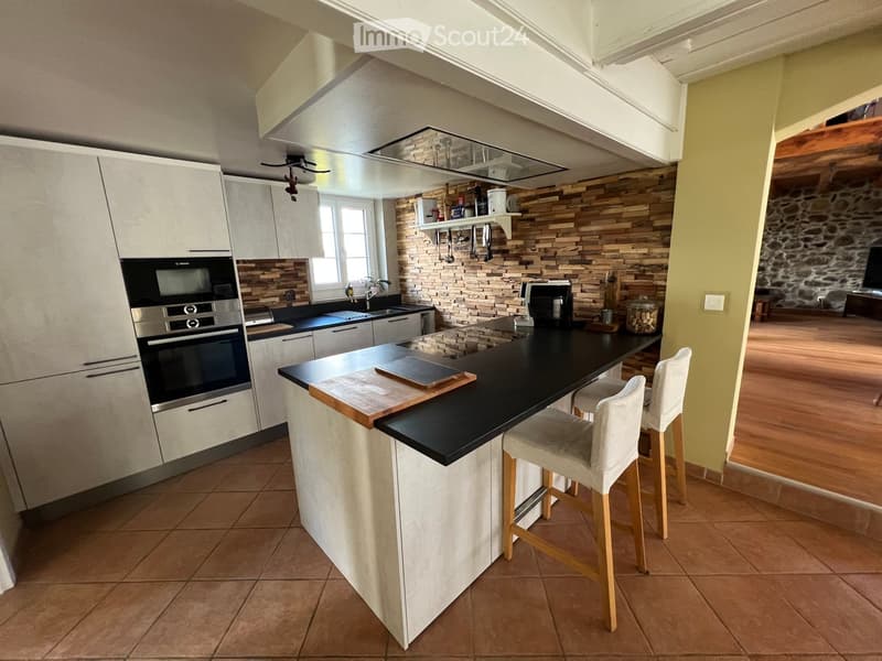 Modern kitchen with white cabinets, black countertops, a central island, and a built-in oven. The space features a wooden backsplash and includes bar stools, a sink, and various appliances.