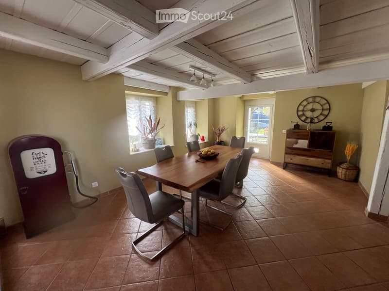Dining room with a wooden dining table, six chairs, a large wall clock, a wooden cabinet, and potted plants near the windows.