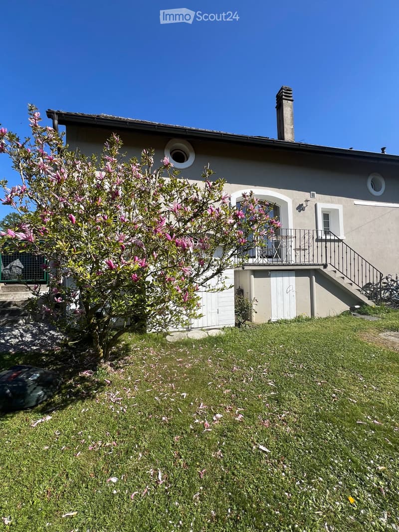Two-story house with beige walls, white-framed windows, and a small balcony on the upper level. A flowering tree is in the front yard, and a raised ground floor is visible.