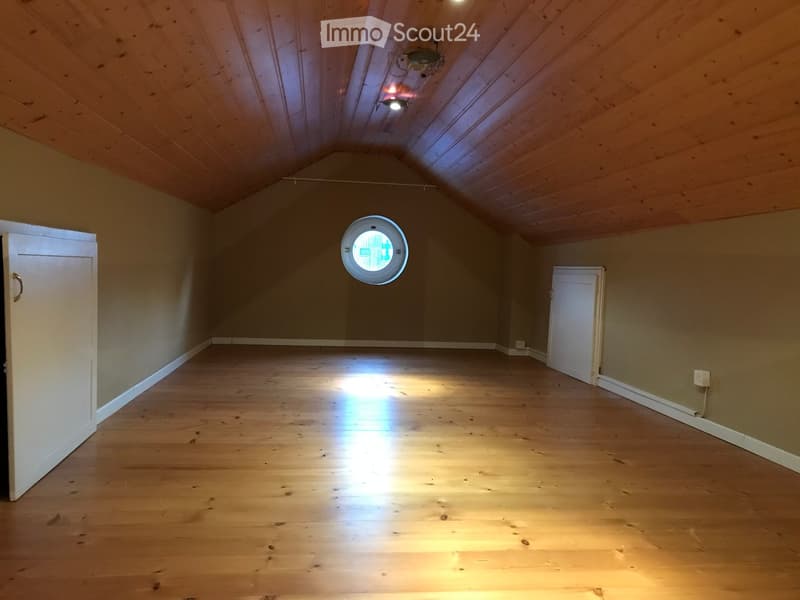 Empty attic room with wooden floors, sloped wooden ceiling, and a round skylight window. Two white doors are visible on either side.