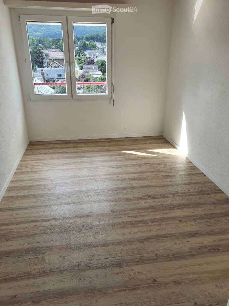 Unfurnished Room. An empty room with wooden flooring and white walls. There are two large windows with a view of the outside. The room is illuminated by sunlight coming from the windows.