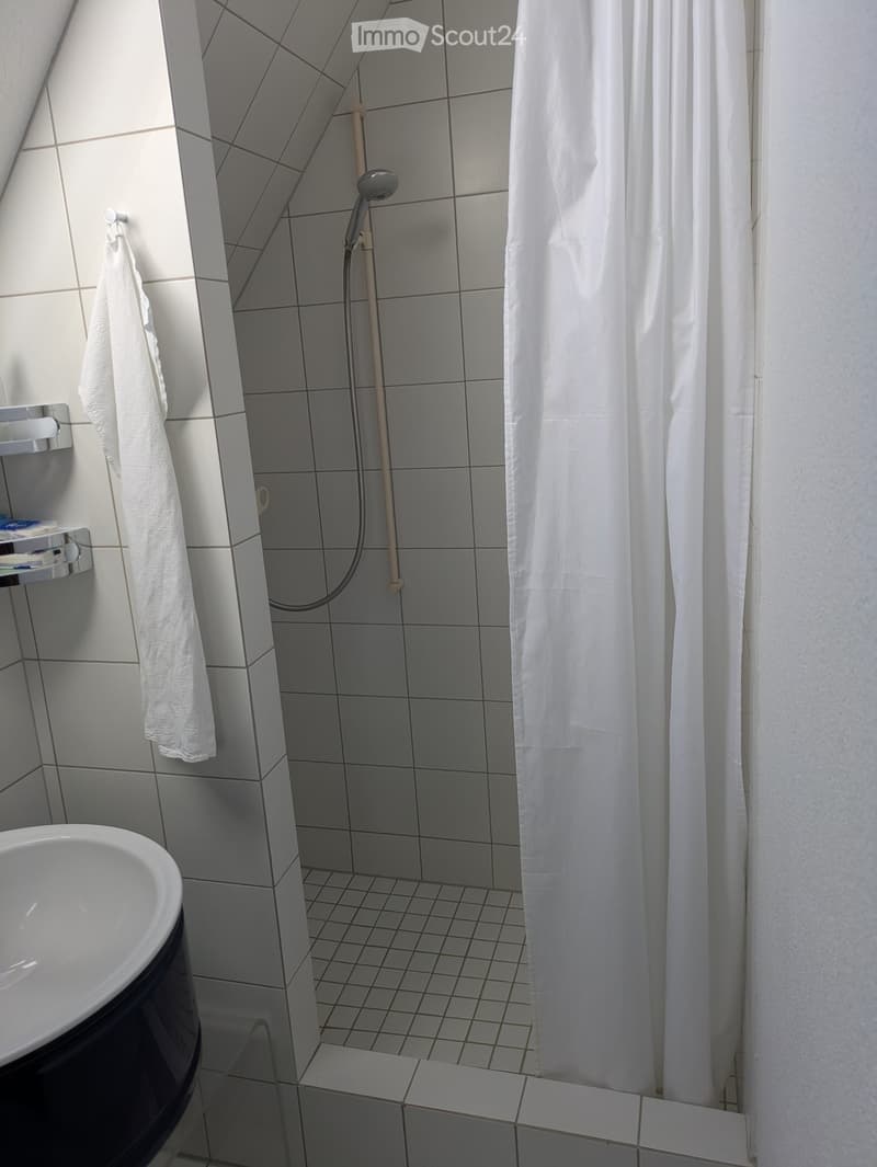 A small bathroom with a shower, sink, and towel rack. The shower is equipped with a shower head and a white shower curtain. The sink is white with a black basin. A towel is hanging on the towel rack.