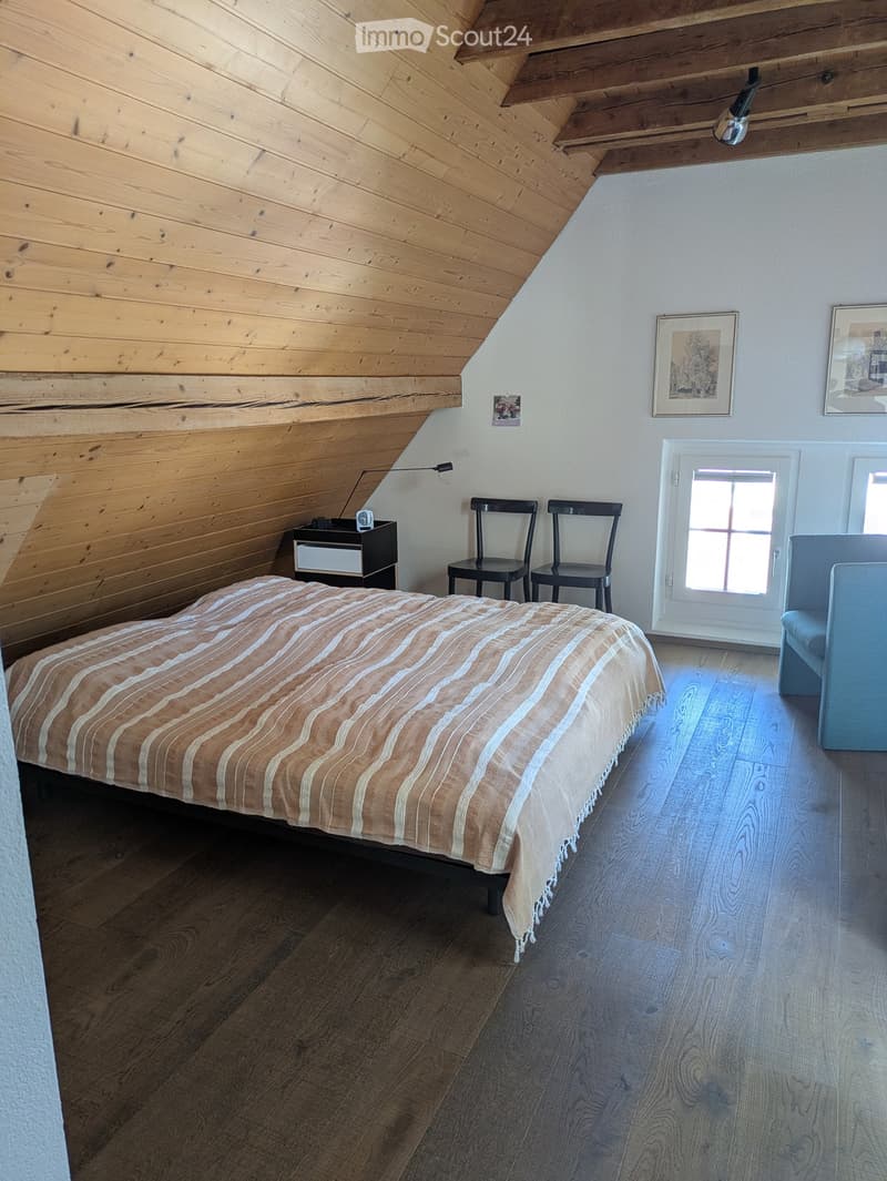 A bedroom with wooden ceiling, bed, nightstand, lamp, two chairs, and a couch. The room has a white wall and wooden floor.