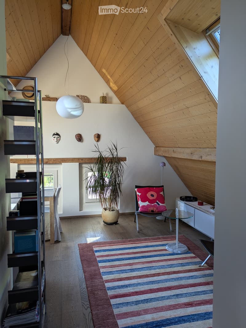 A cozy attic room with wooden ceiling, a bookshelf, a chair, a potted plant, a rug, a lamp, a table, and a side table.