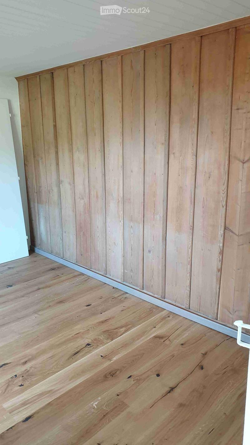 An empty room with wooden walls and a wooden floor. The walls are made of vertical wooden panels, and the floor is covered with wooden planks. There is a white door on the left side of the room.