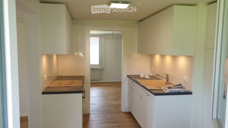 Modern kitchen with white cabinets, wooden floor, and a sink with a faucet. Countertops are empty with some paper on them.