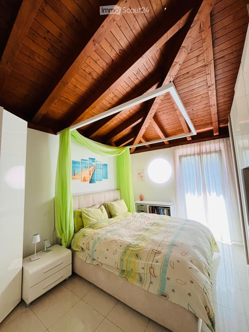 Bedroom with wooden ceiling, bed, nightstand, lamp, bedspread, pillows, curtains, paintings, shelf, books, and sculpture.