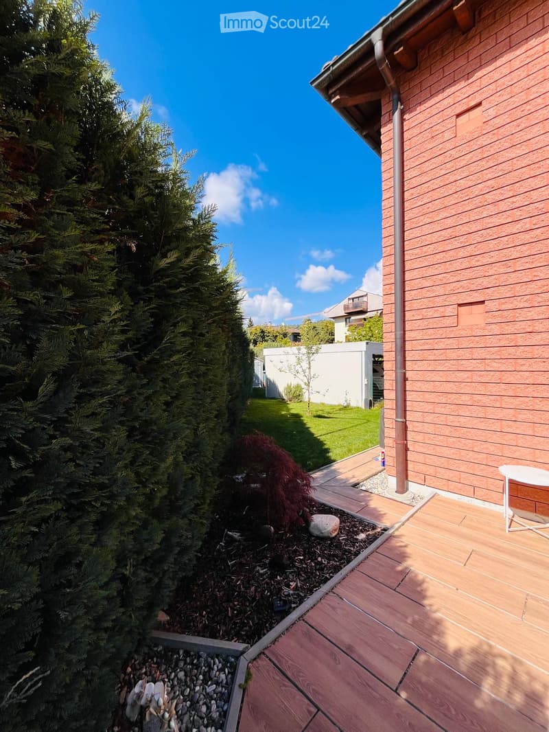 A wooden deck with a small garden area featuring a hedge, a small plant, and a rock. The garden is bordered by a red brick wall.