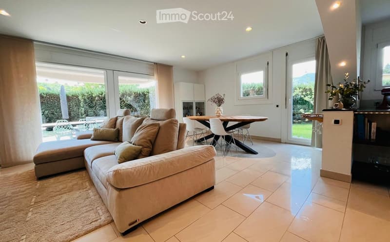 Spacious living room with beige sofa, coffee table, dining table, and chairs. Large windows with curtains. Beige rug and decorative items.