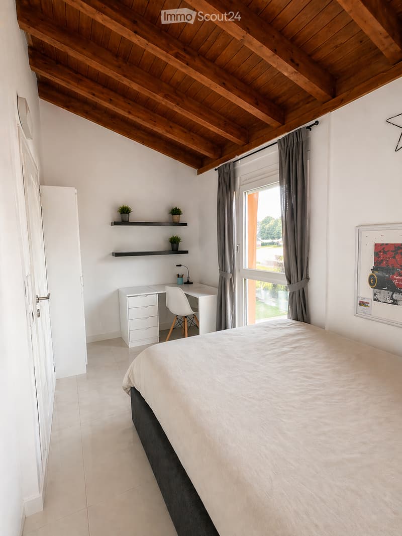 A bedroom with a bed, desk, chair, lamp, plants, cabinet, and a framed picture on the wall.