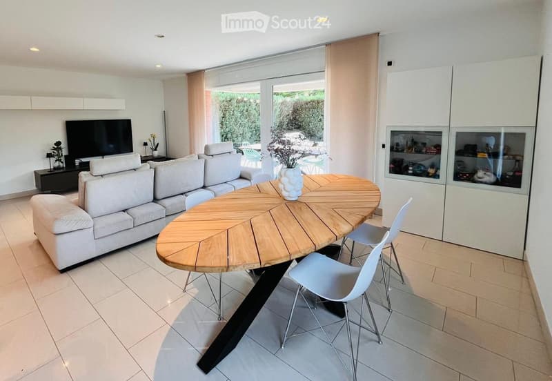 Modern living room with a white couch, wooden dining table, and white chairs. A TV is on a stand, and a vase with flowers is on the table. A cabinet with glass doors is on the right side.
