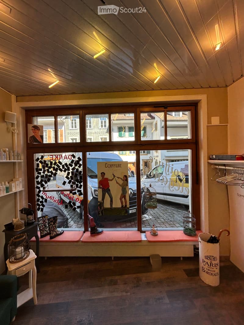 A room with a bench, umbrella stand, and a table with a candle. There is a picture and a poster on the wall. Shoes are placed on the bench. A van and a car are parked outside the window. A lamp is mounted on the wall. A basket, plant, and trash can are on