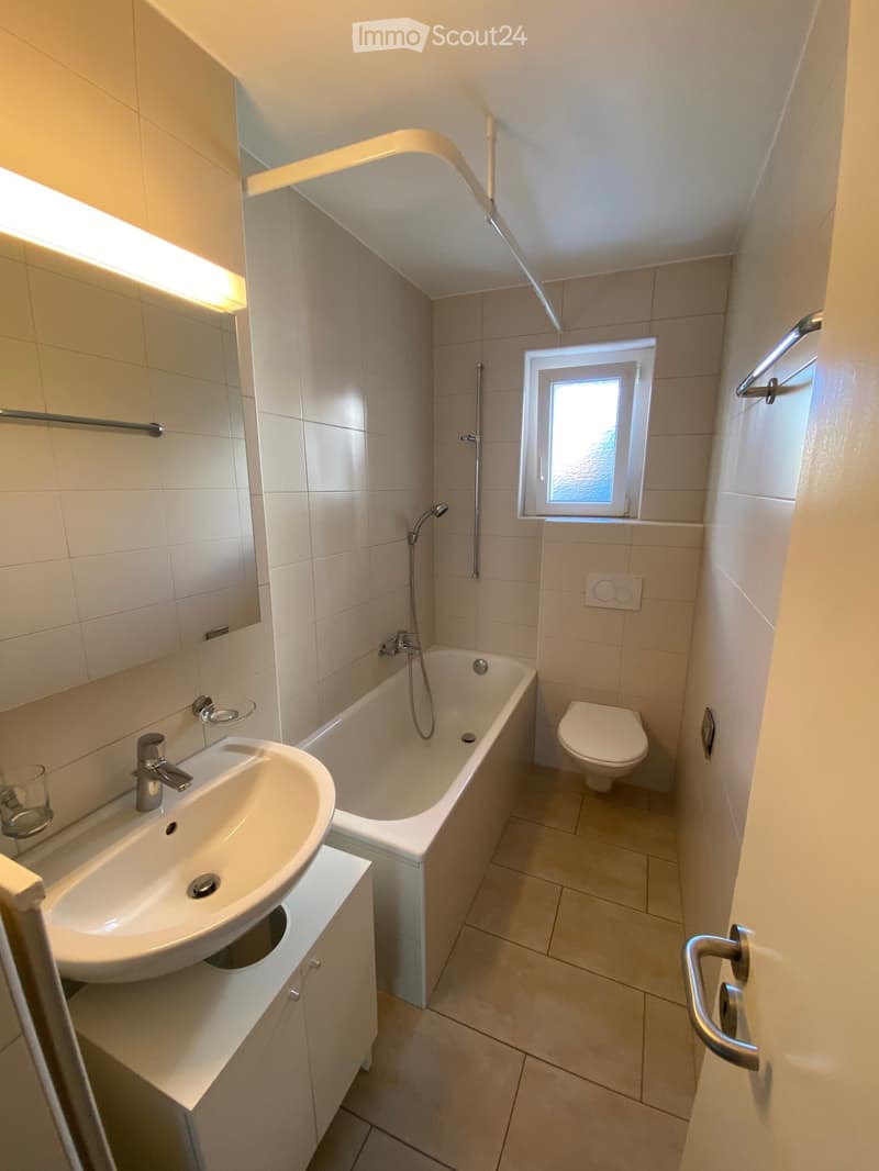 Modern bathroom with a white sink, bathtub, toilet, and a large mirror. The room features beige tiles and a small window letting in natural light.