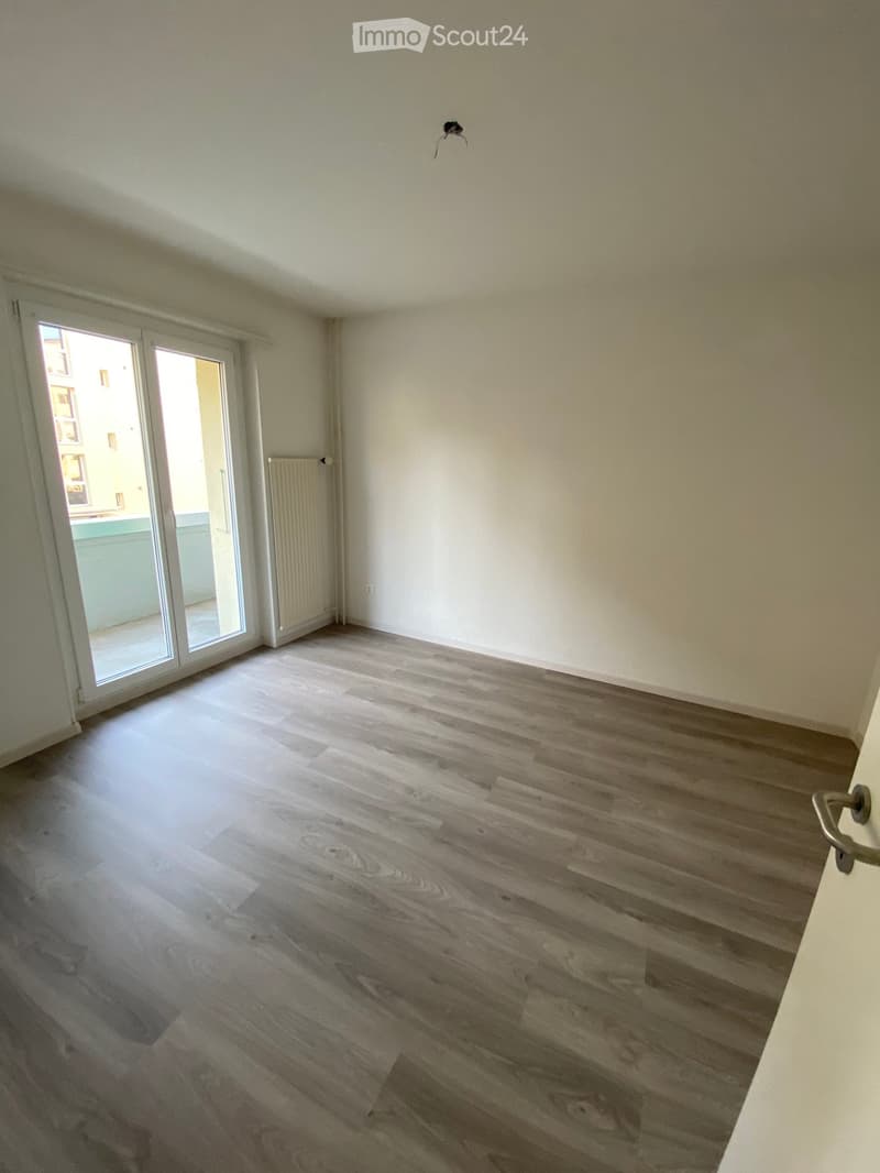 Empty room with wooden flooring, white walls, large sliding glass door leading to a balcony, radiator, and a small light fixture on the ceiling.