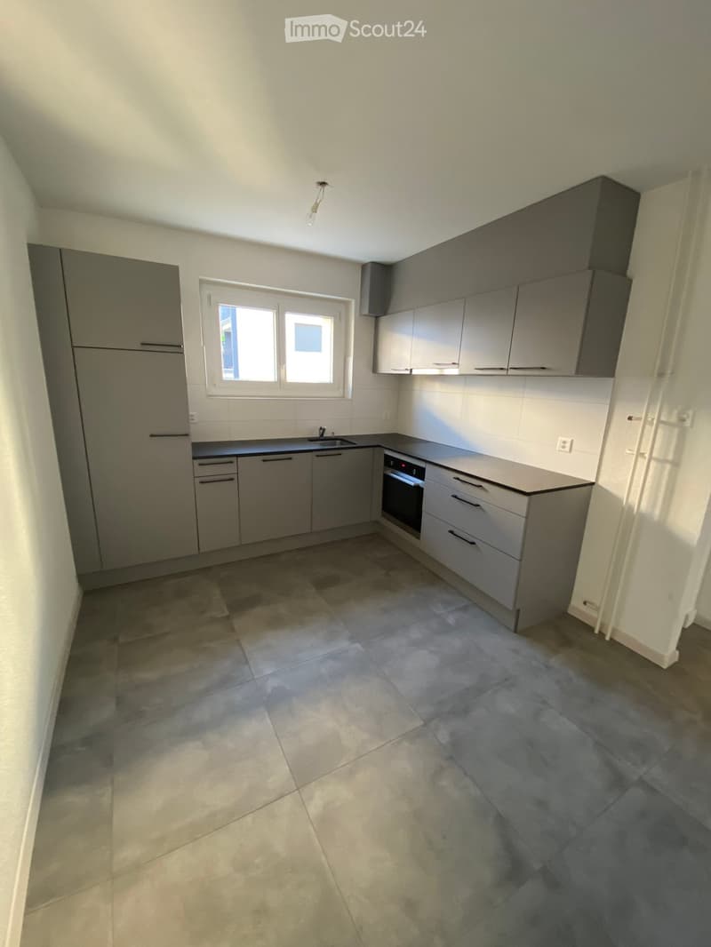 Modern kitchen with grey cabinets, a refrigerator, an oven, and a sink, featuring a tiled floor and a window letting in natural light.