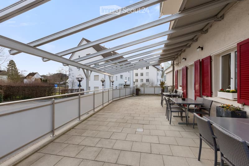 outdoor terrace, tables and chairs, metal railing, covered area, greenery in background