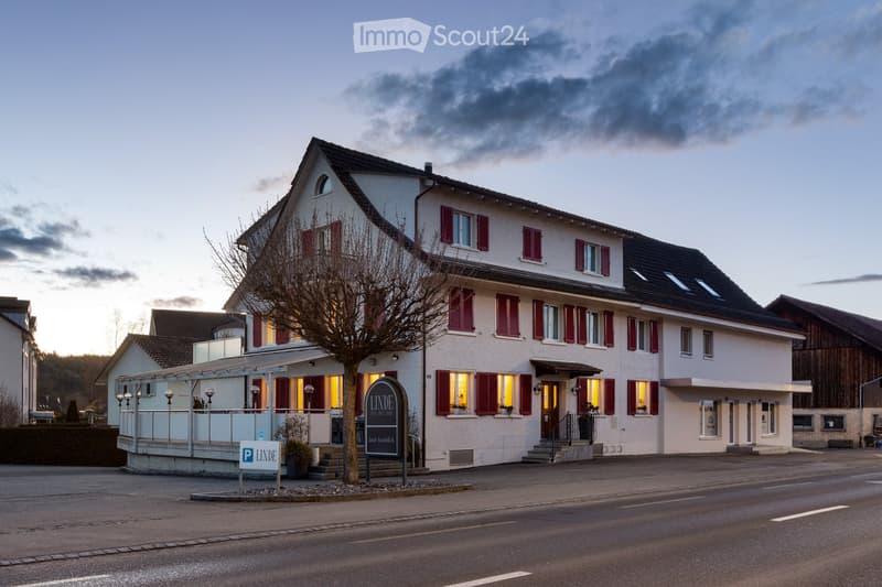 Building: white building, red shutters, black roof, glassed front with stairs, signage 'LINDE Weinhaus', balcony on the left, parking in front
