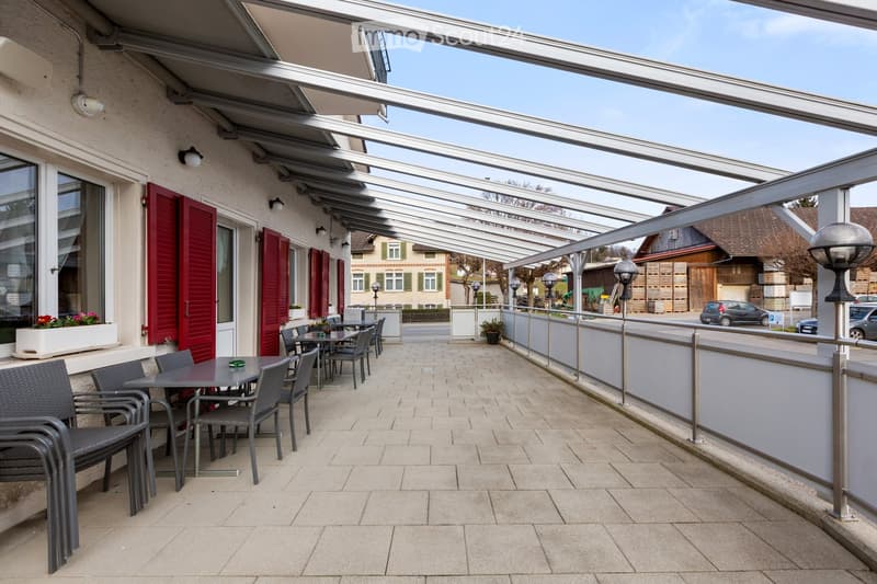Tiled floor, metal railing, chairs, tables, transparent canopy, red shutters, street view