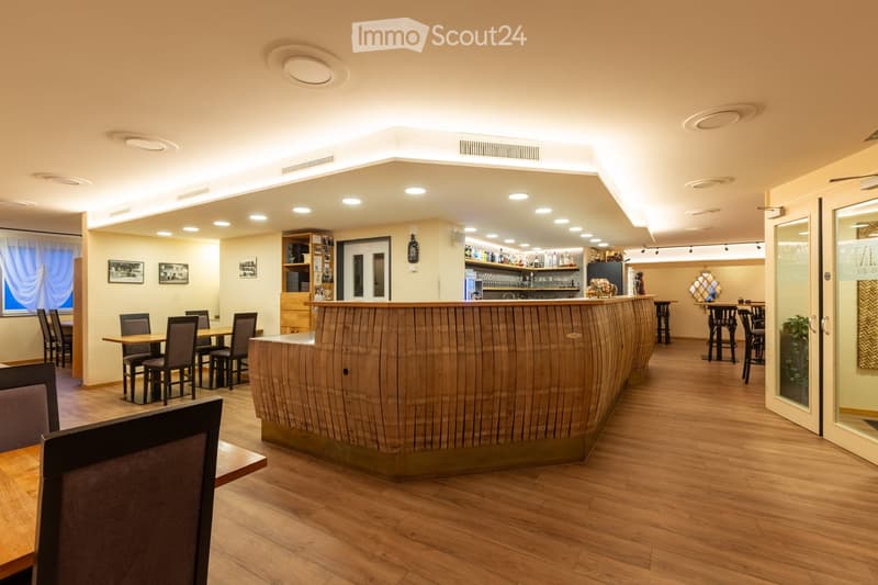 interior of a restaurant with wooden flooring, wooden reception desk, wooden tables and chairs, shelves with bottles, and frames on the wall