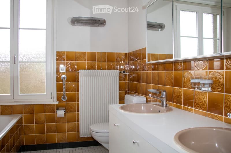 Bathroom with white and brown tiles, white bathtub, white toilet, double sinks, and a mirror.