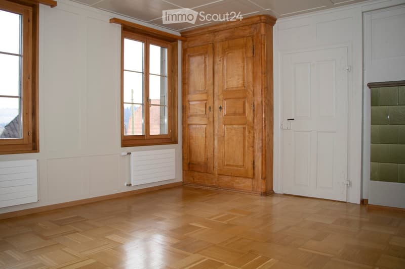 Empty room with wooden parquet flooring, white walls, wooden window frames, and a large wooden cabinet.