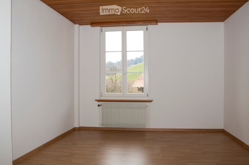 Empty room with white walls, wooden ceiling, and wooden floor. A window and a radiator are present.