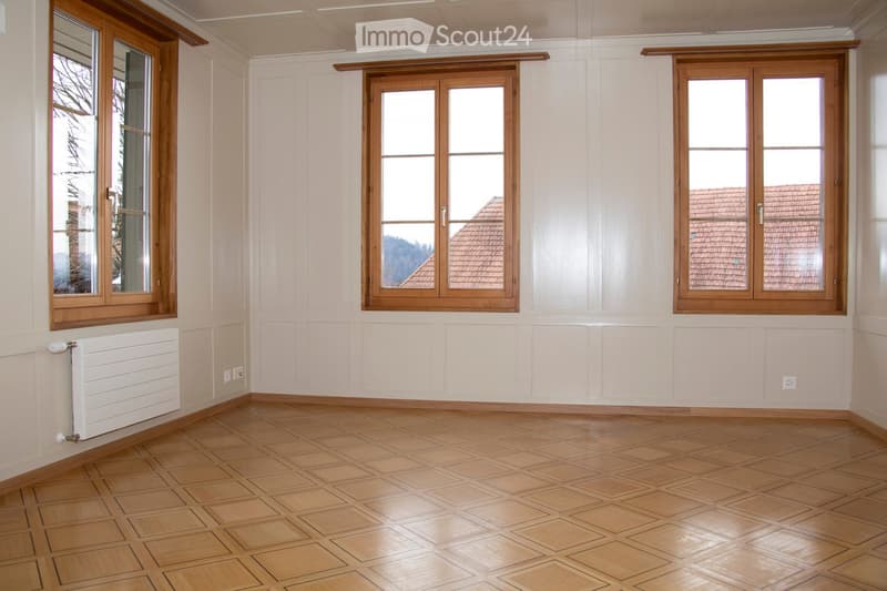 Empty room with wooden parquet flooring, white walls, and three large windows with wooden frames.