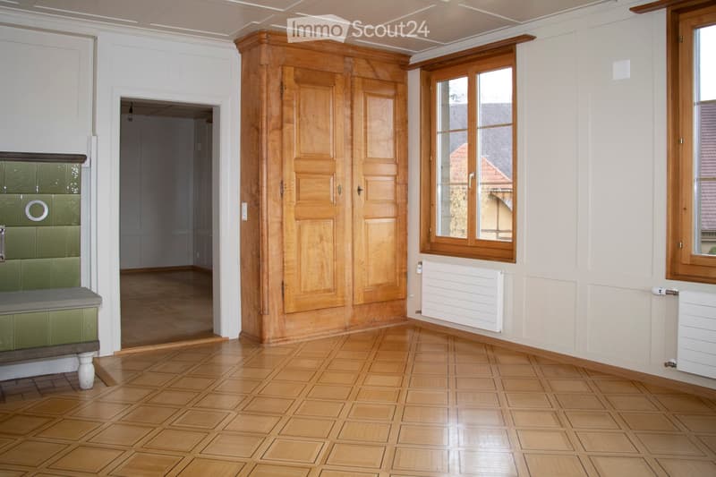 Empty room with wooden parquet flooring, wooden wardrobe, white walls, and green tiled area. Two windows with wooden frames and white radiators.