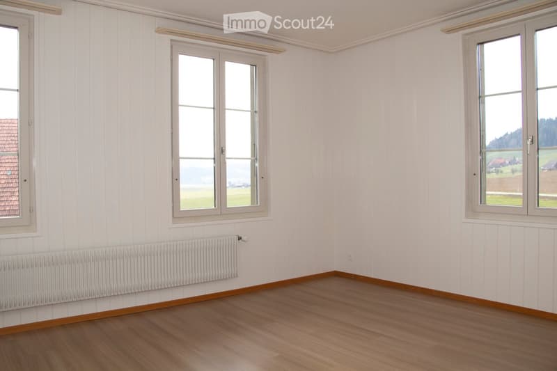 Empty room with white walls, wooden floor, and three windows. There is a white radiator on the left wall.