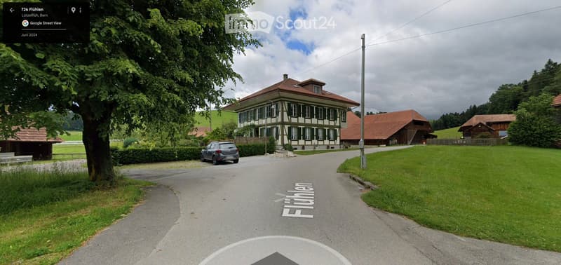 A street view of a house with a car parked in front. The house has a brown roof and white walls with green shutters. The house is surrounded by green grass and bushes. There are other houses in the distance, and the sky is cloudy.