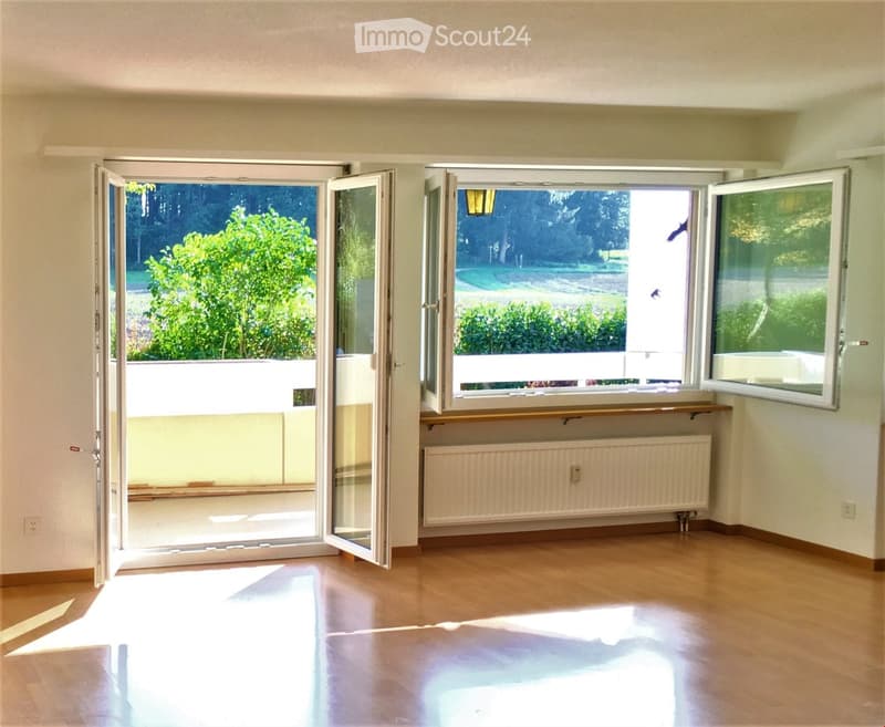 Empty room with wooden floors, white walls, and open sliding glass doors leading to a balcony. A radiator is mounted on the wall below the windows.