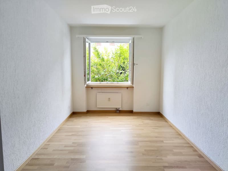 Empty room with light wooden flooring, white walls, and a large window with a view of greenery. A radiator is mounted on the wall below the window.