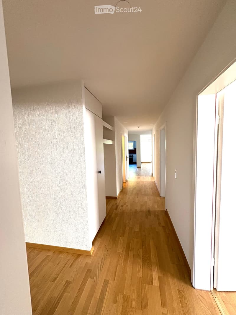 Empty hallway with light wooden flooring and white walls, featuring several doors and a visible room at the end.